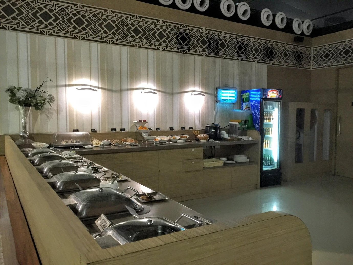 Buffet area in the T1 travel club lounge at Kolkata airport, featuring a variety of hot food dishes, snacks, beverages, and a refrigerator stocked with drinks, set in a modern interior with intricate wall decor.