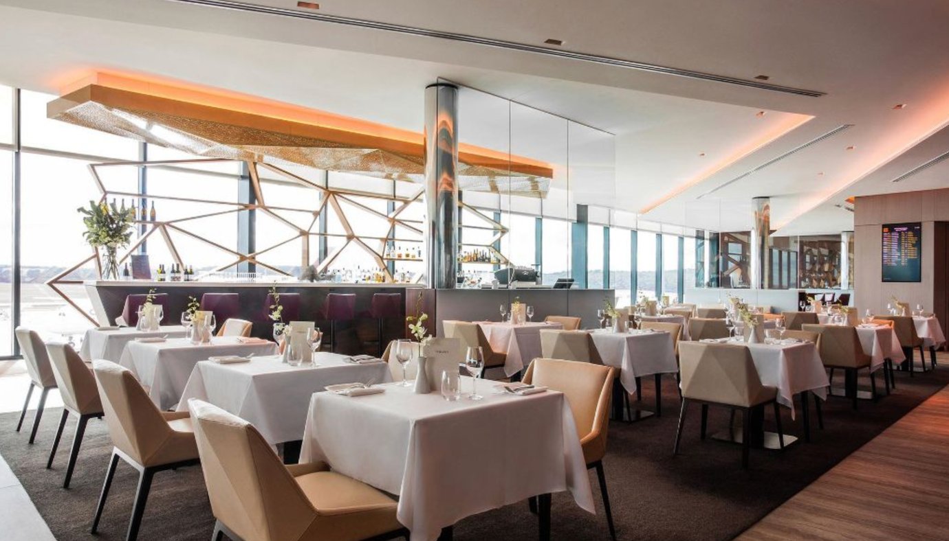 Aspire Lounge dining area at Melbourne Airport with white tablecloth seating, warm lighting, and runway views.