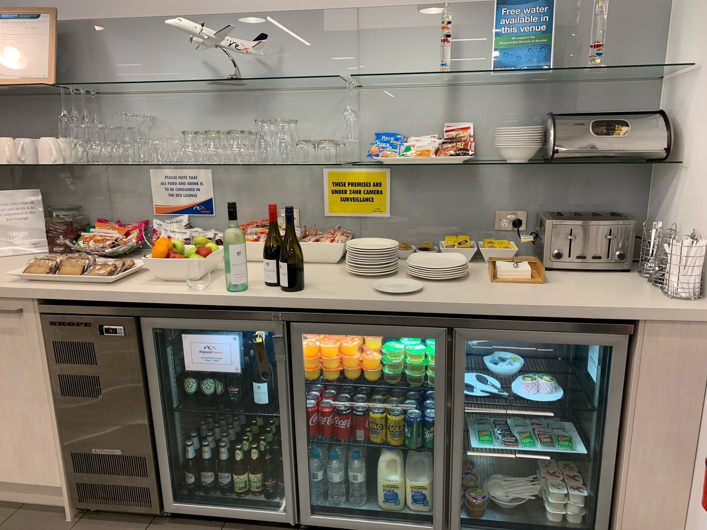 Rex Lounge at Melbourne Airport self-service refreshment area with snacks, fruit, wine, and refrigerated drinks on display. Rex Lounge at Melbourne Airport self-service refreshment area with snacks, fruit, wine, and refrigerated drinks on display.