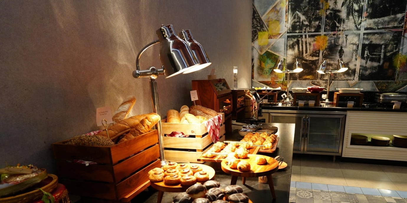 Breakfast buffet at Concordia Lounge, Ngurah Rai International Airport (DPS) Bali, featuring fresh pastries, bread baskets, croissants, and hot dishes under warming lamps.