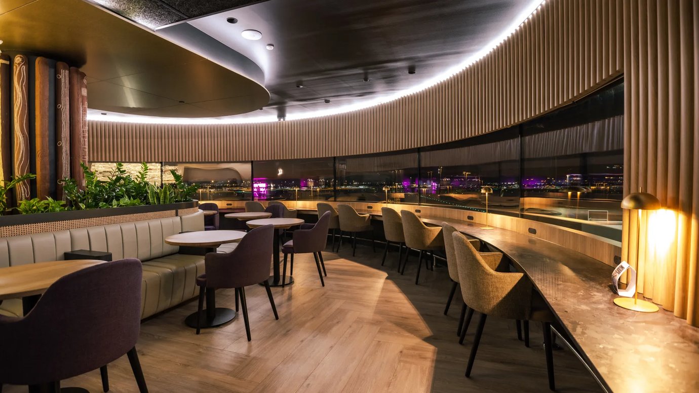 Aspire Lounge Perth seating area with curved window-side workstations, upholstered dining chairs, and booth seating overlooking the airport apron at night.