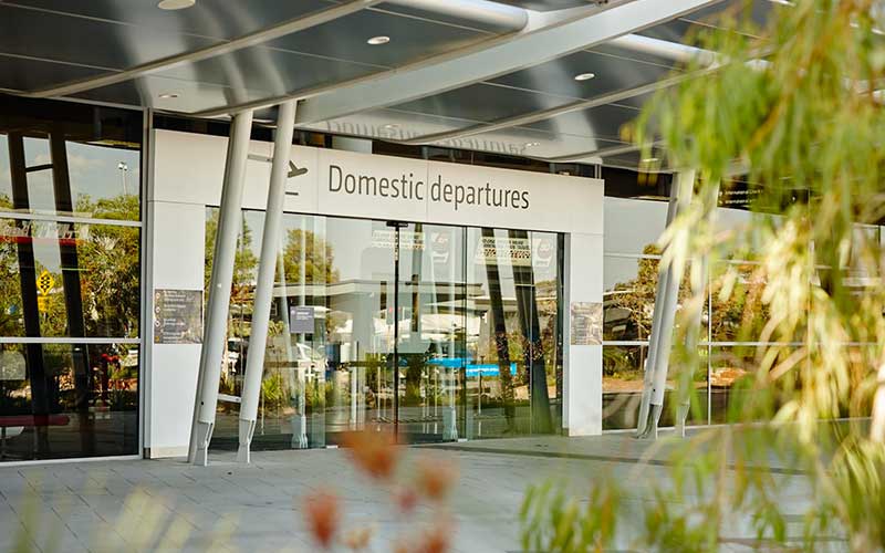 Perth Airport domestic departures entrance with sliding glass doors, overhead terminal signage, and a covered exterior walkway supported by angled steel columns.