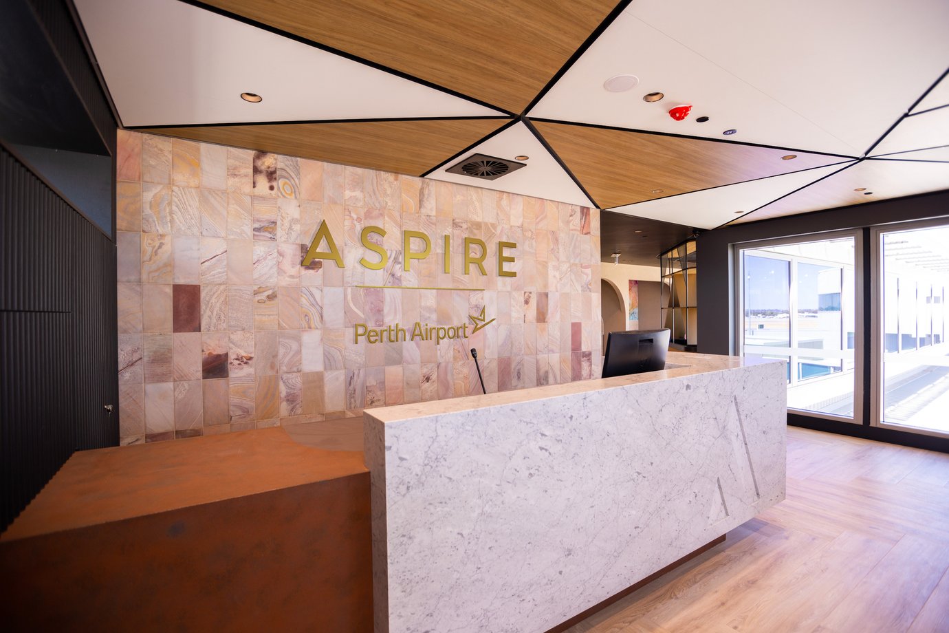 Aspire Lounge Perth Airport reception area with a marble check-in desk, tiled feature wall with Aspire branding, and geometric wood-panel ceiling design.
