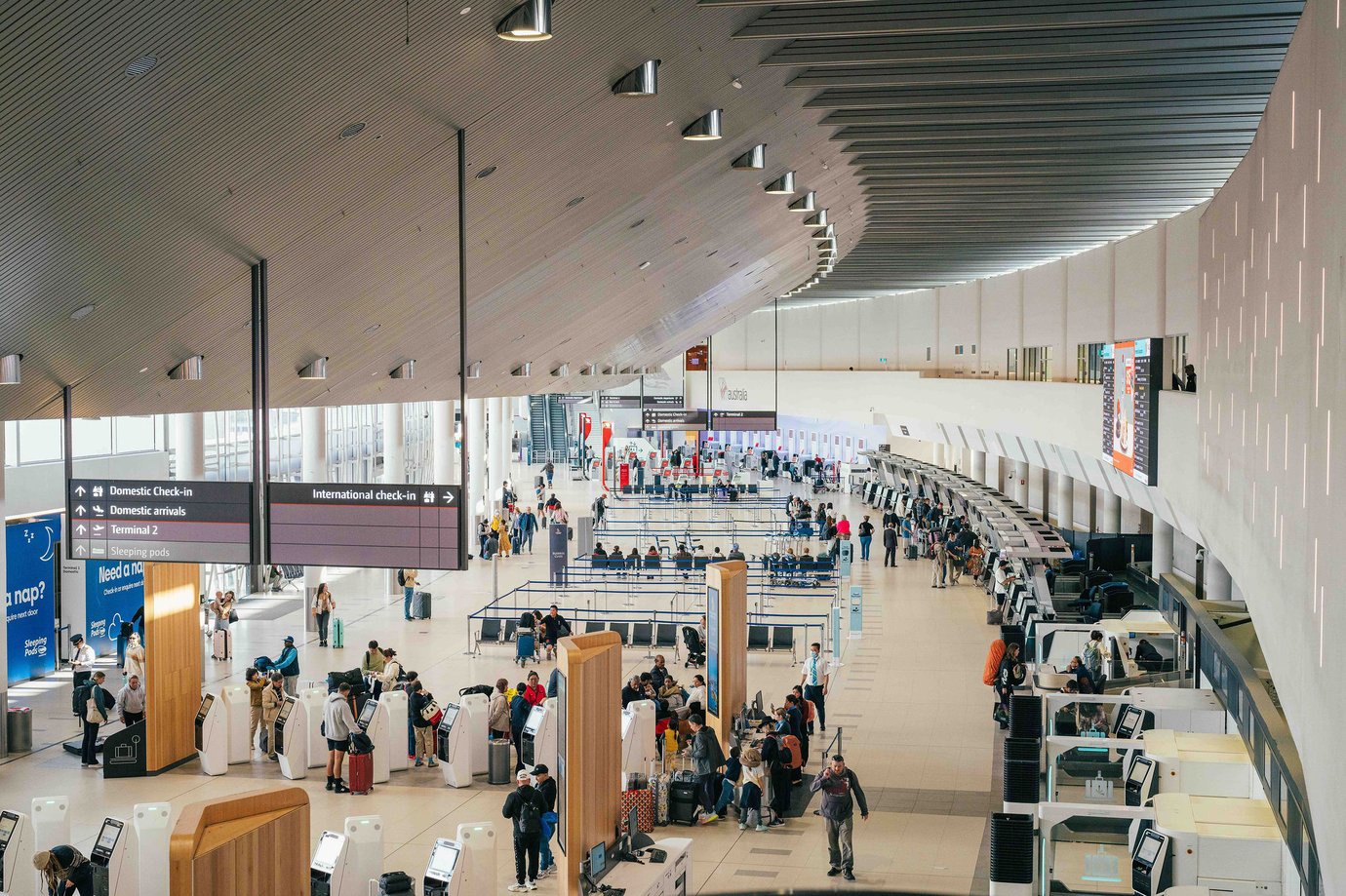 Perth Airport check-in hall with self-service kiosks, airline counters, and passenger queue areas beneath a curved timber-style ceiling.