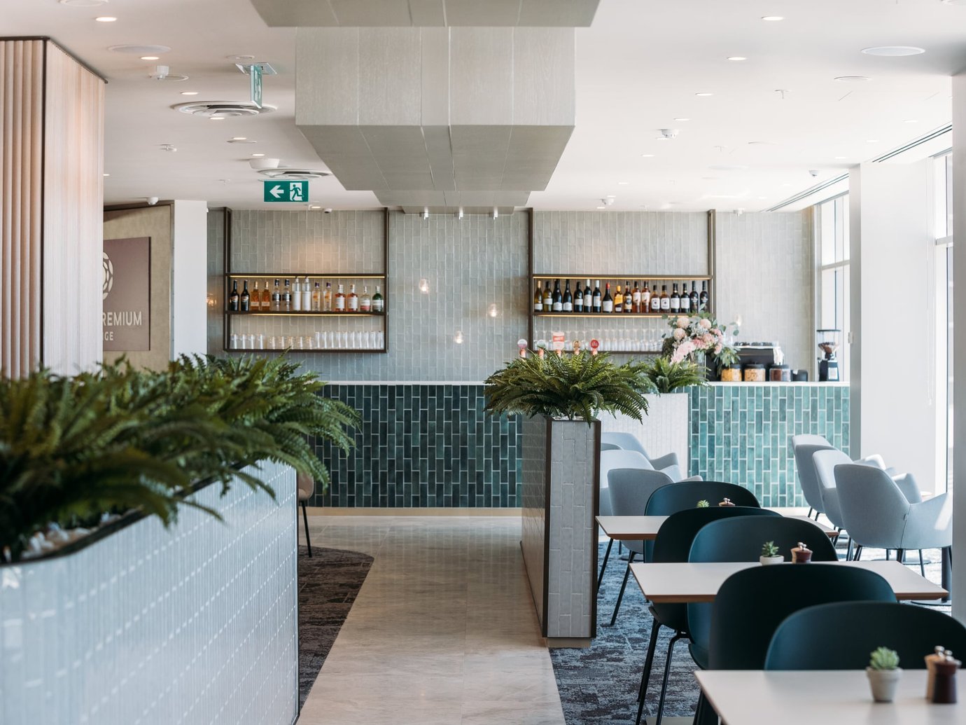 Plaza Premium Lounge at Sydney Airport bar area with tiled counter, backlit shelves of wine and spirits, and dining tables with lounge seating.