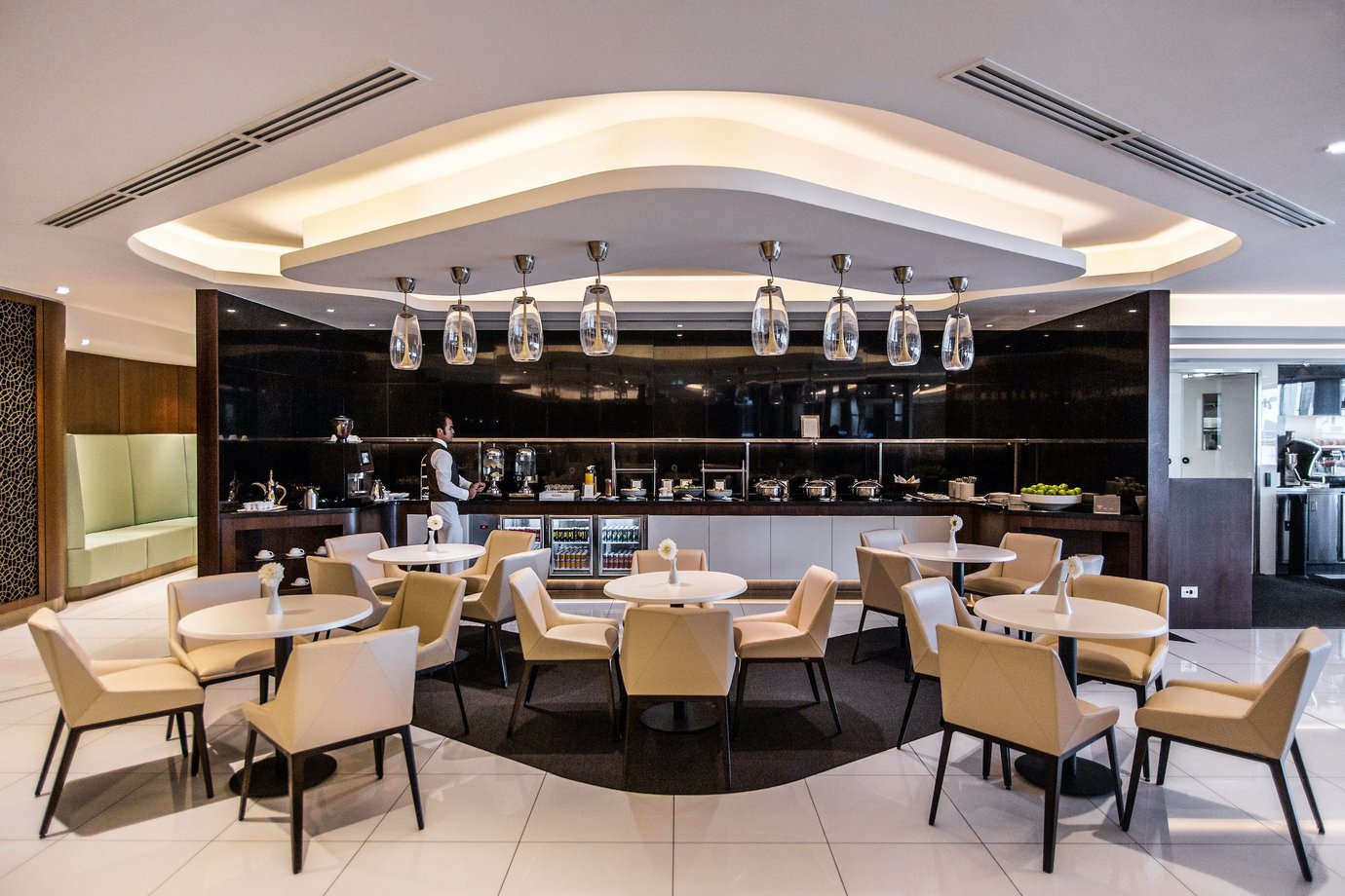 The House lounge at Sydney Airport, featuring a central bar with a bartender, warm ambient lighting, and small round tables with beige chairs arranged across the seating area.