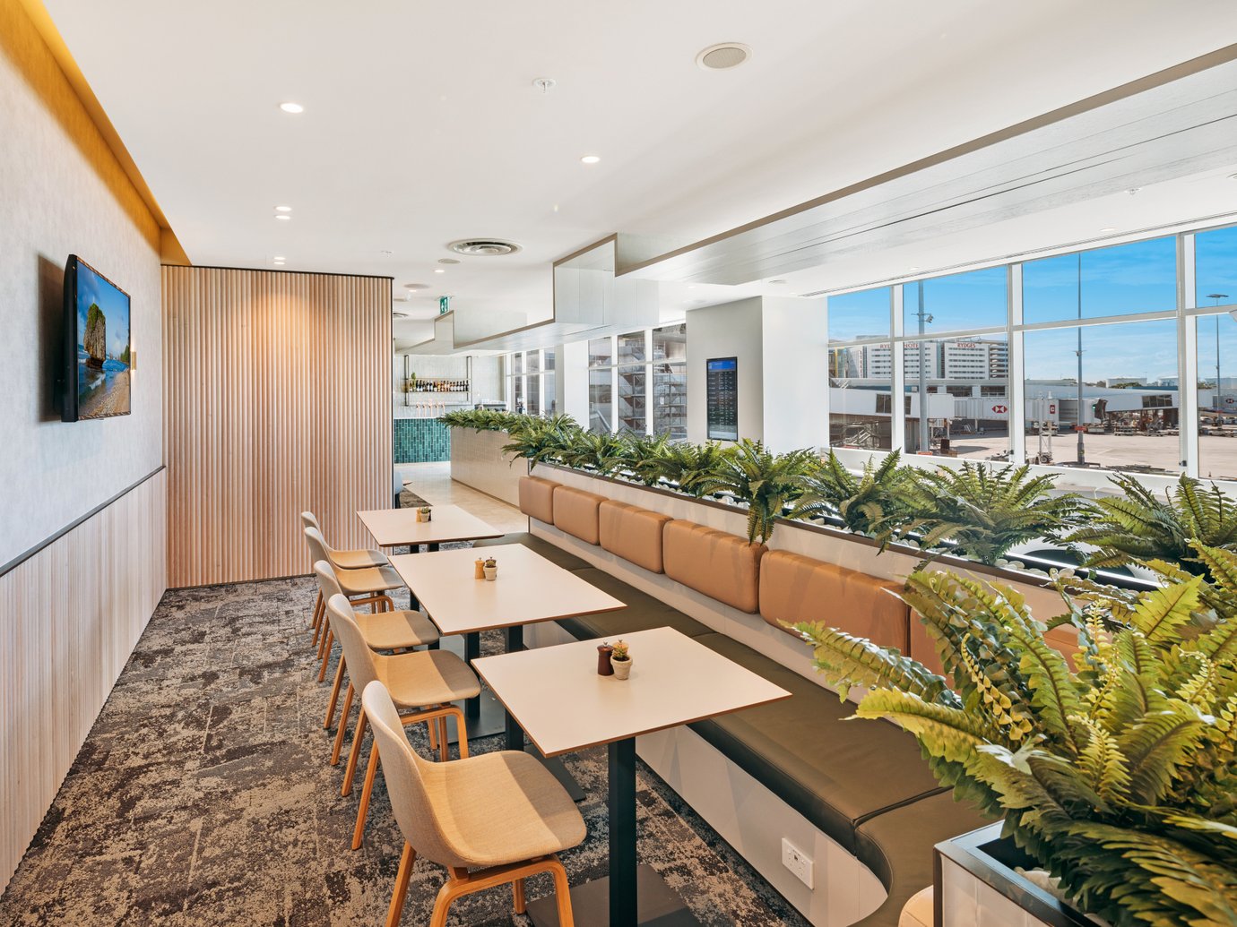 Plaza Premium Lounge at Sydney Airport departures area, window-side seating with tables, cushioned bench seating, and views of the airport apron.