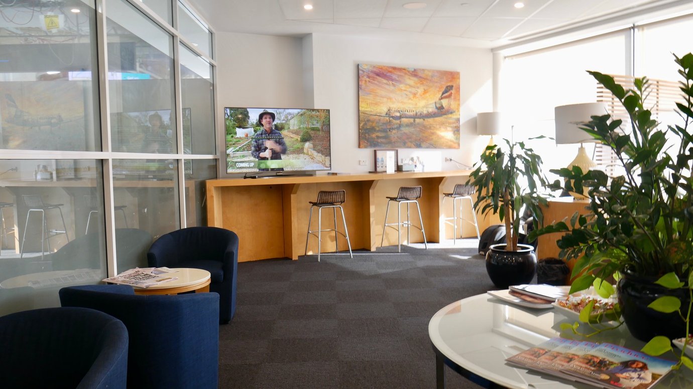 REX Lounge at Sydney Airport seating area with armchairs, TV viewing space, bar-style seating, and natural light from large windows.