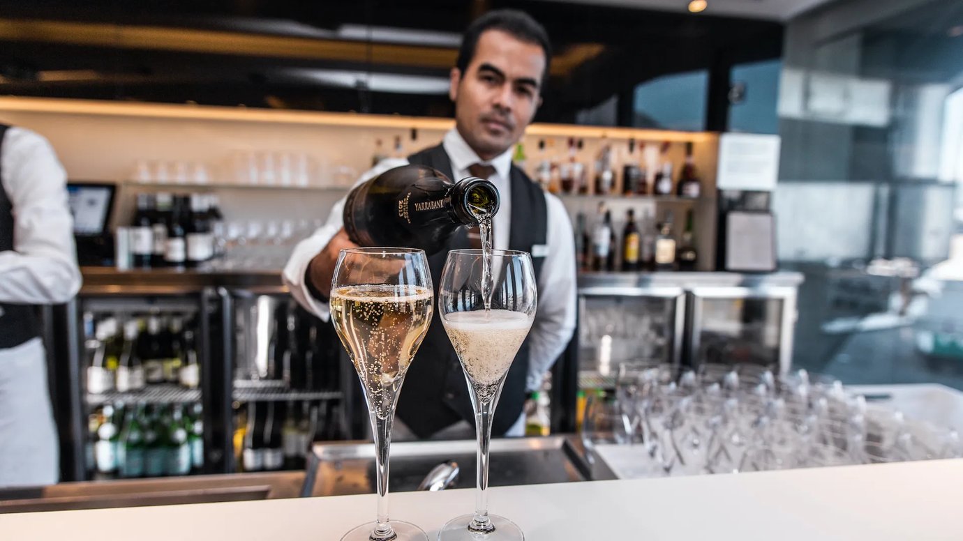 The House lounge at Sydney Airport, bartender pouring sparkling wine into glasses at the bar with bottles and glassware in the background.