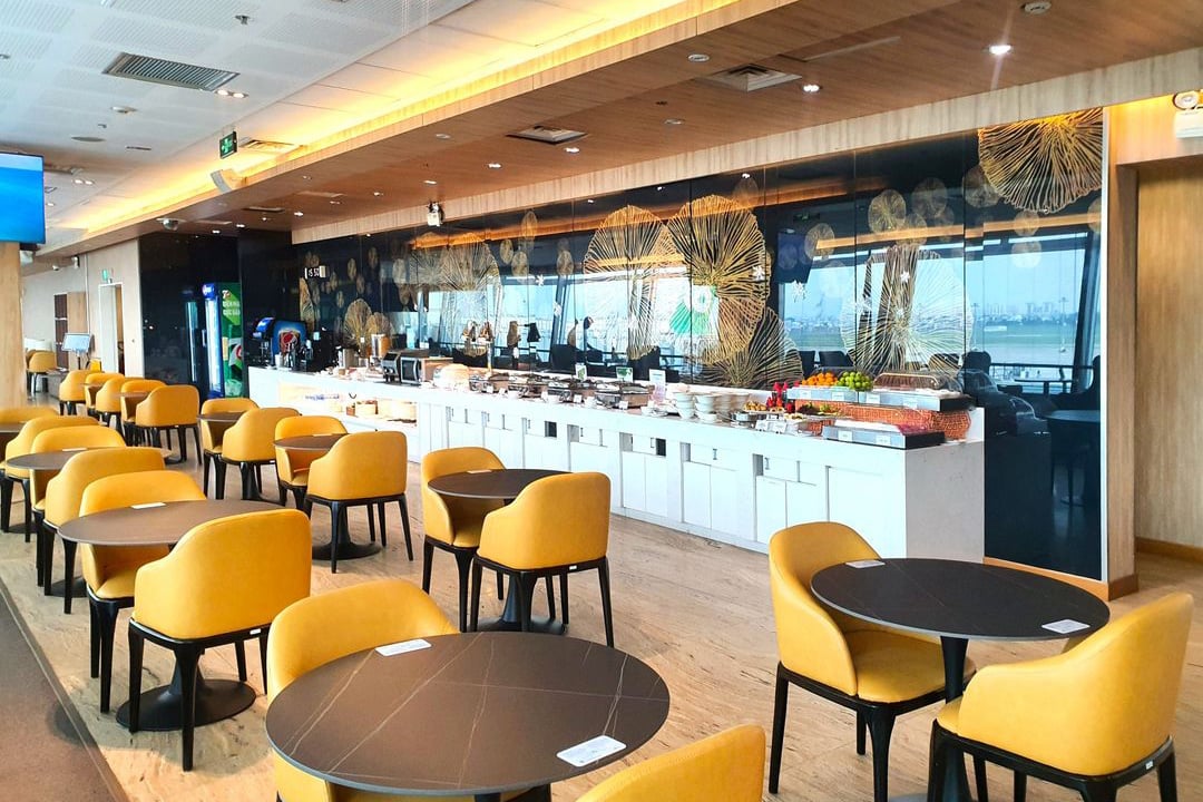 Dining area of the Apricot Business Lounge at Tan Son Nhat Airport, showing rows of mustard-yellow chairs and round tables facing a long self-serve buffet