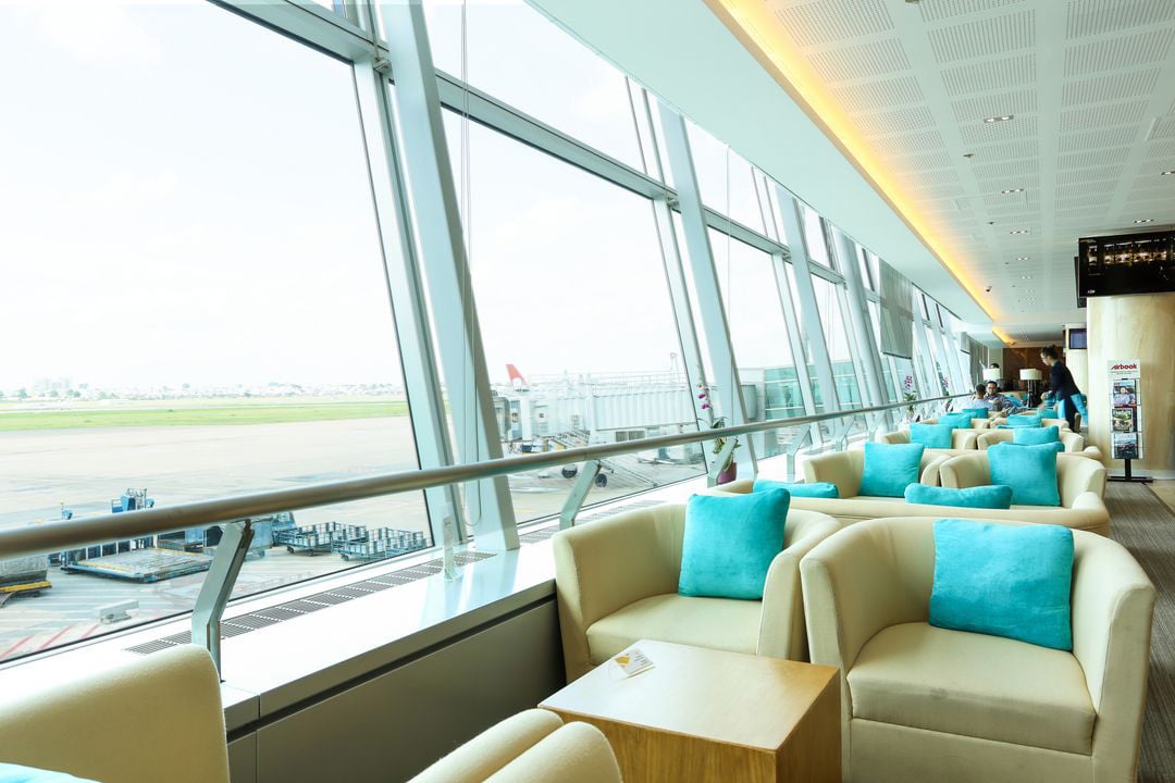 Comfortable sofa seating inside the Apricot Business Lounge at Tan Son Nhat Airport, featuring cream armchairs with teal cushions positioned beside floor-to-ceiling windows overlooking the airport runway.