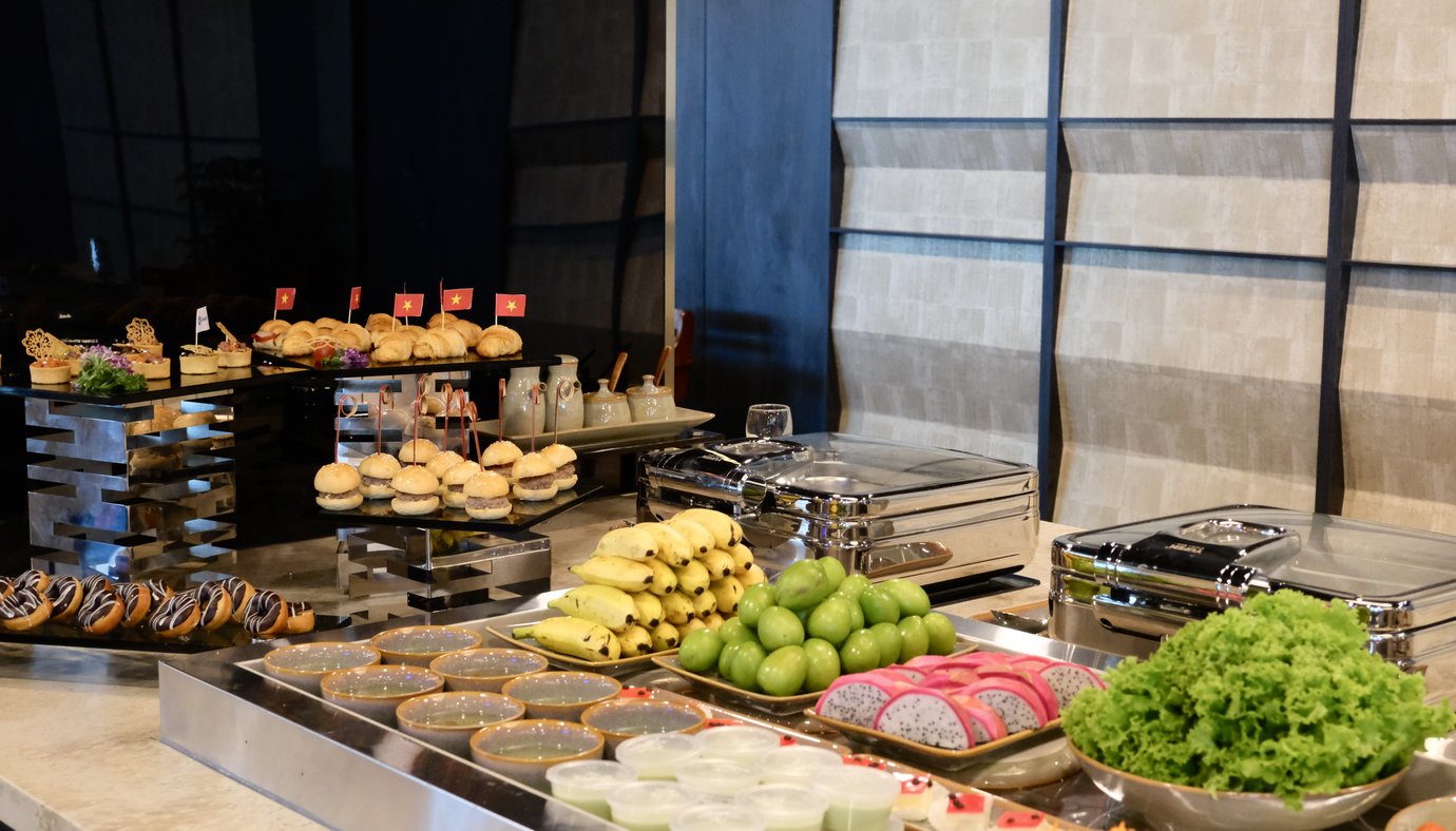 Buffet spread at The Sens Business Class Lounge, Tan Son Nhat Airport, featuring fresh fruit (bananas, green apples, dragon fruit), leafy greens, covered hot dishes, and an array of pastries and mini sandwiches displayed on modern tiered stands