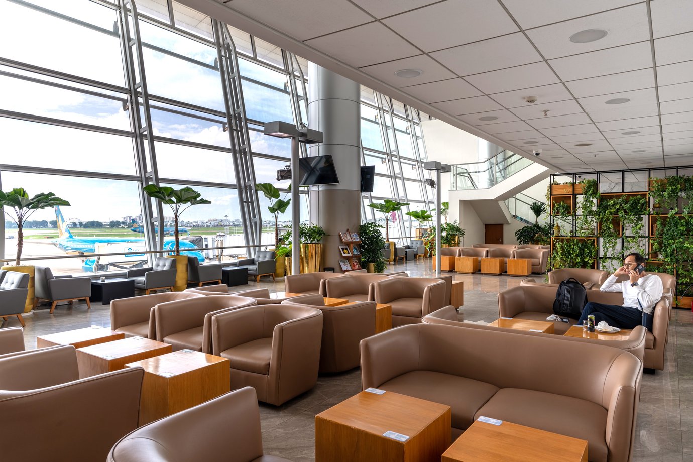 Bright airport lounge with tan leather sofas, wood side tables, indoor plants, and floor-to-ceiling windows overlooking the runway.