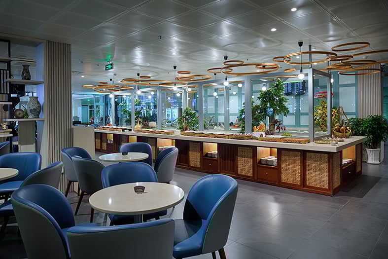 Buffet area inside Le Saigonnais Business Lounge at Tan Son Nhat Airport, featuring a long wooden buffet counter, hanging circular light fixtures, indoor plants, and blue lounge chairs with round tables.