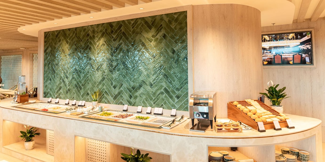 Buffet counter inside the SH Premium Lounge at Tan Son Nhat Airport, featuring a curved stone island with chilled salad trays, bread and roll display, toaster station, and small potted plants, set against a green herringbone-tiled accent wall