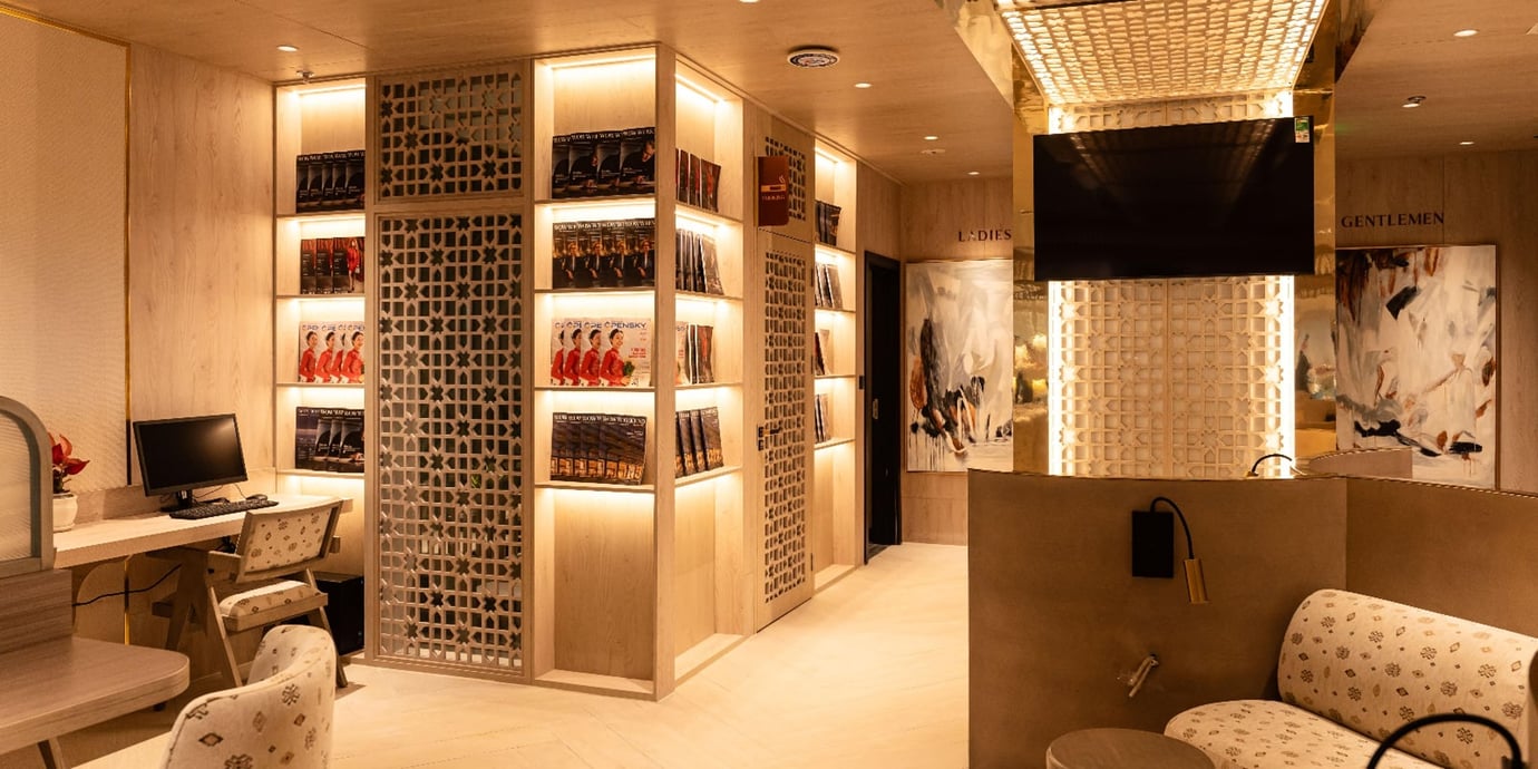 Interior view of the SH Premium Lounge at Tan Son Nhat Airport showing a refined, gold-toned space with decorative lattice panels, illuminated magazine shelves, a small work desk with computer, and clearly marked restrooms