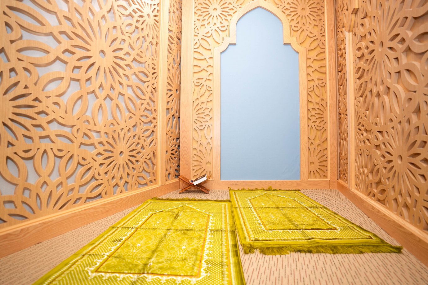 Quiet prayer room inside the Jasmine Halal Lounge at Tan Son Nhat International Airport, featuring ornate carved wooden panels, a mihrab-style niche, two green prayer mats laid side by side, and a Quran placed on a wooden stand