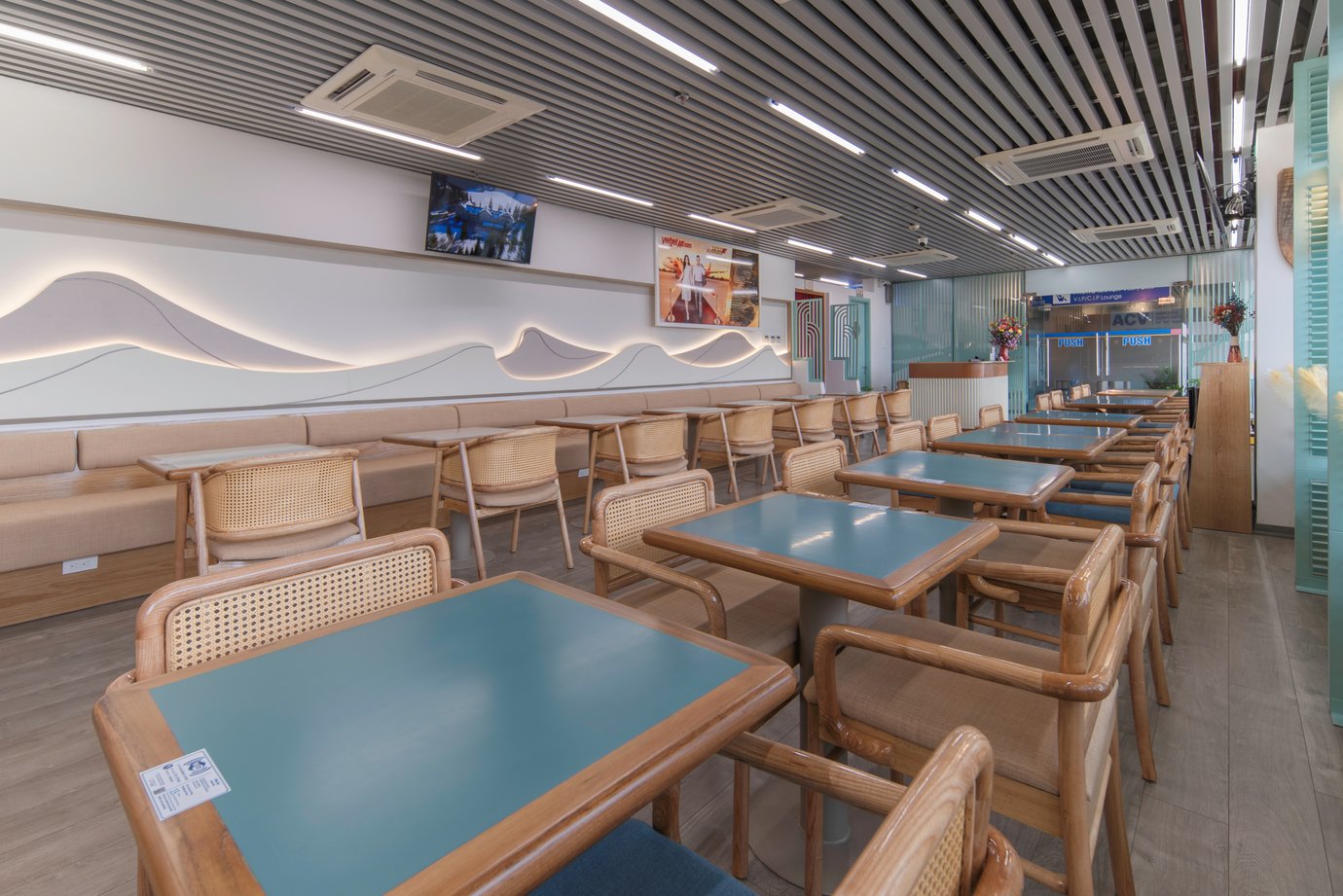 H Premium Lounge at TBB Airport with a long dining room of teal-topped tables and cane-back wooden chairs, a beige banquette along the wall under a wavy LED light feature, and a slatted ceiling H Premium Lounge at TBB Airport with a long dining room of teal-topped tables and cane-back wooden chairs, a beige banquette along the wall under a wavy LED light feature, and a slatted ceiling