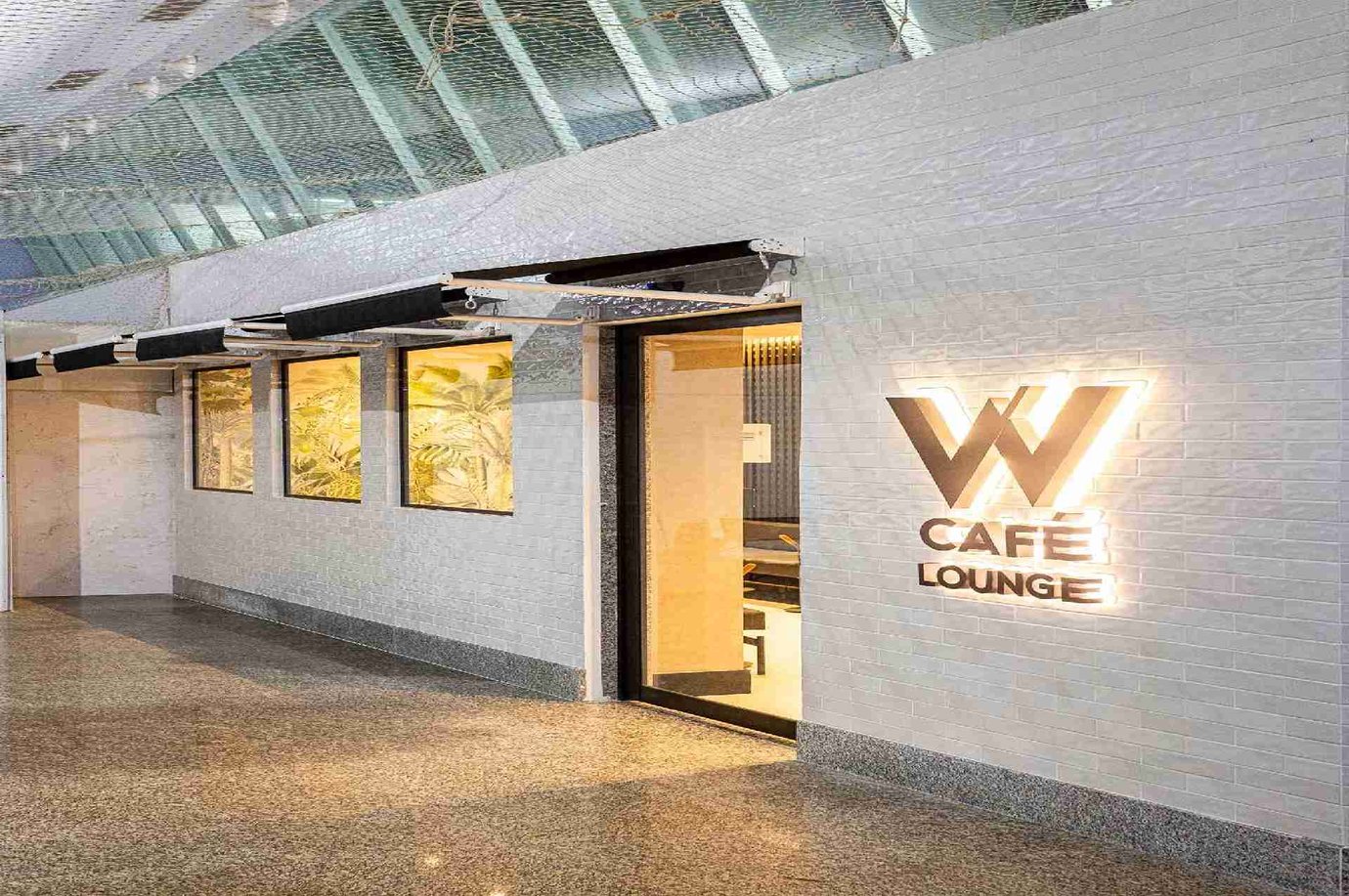 Exterior entrance of W Café Lounge at Palmas Airport, featuring illuminated signage on a white tiled wall and modern glass canopy design.