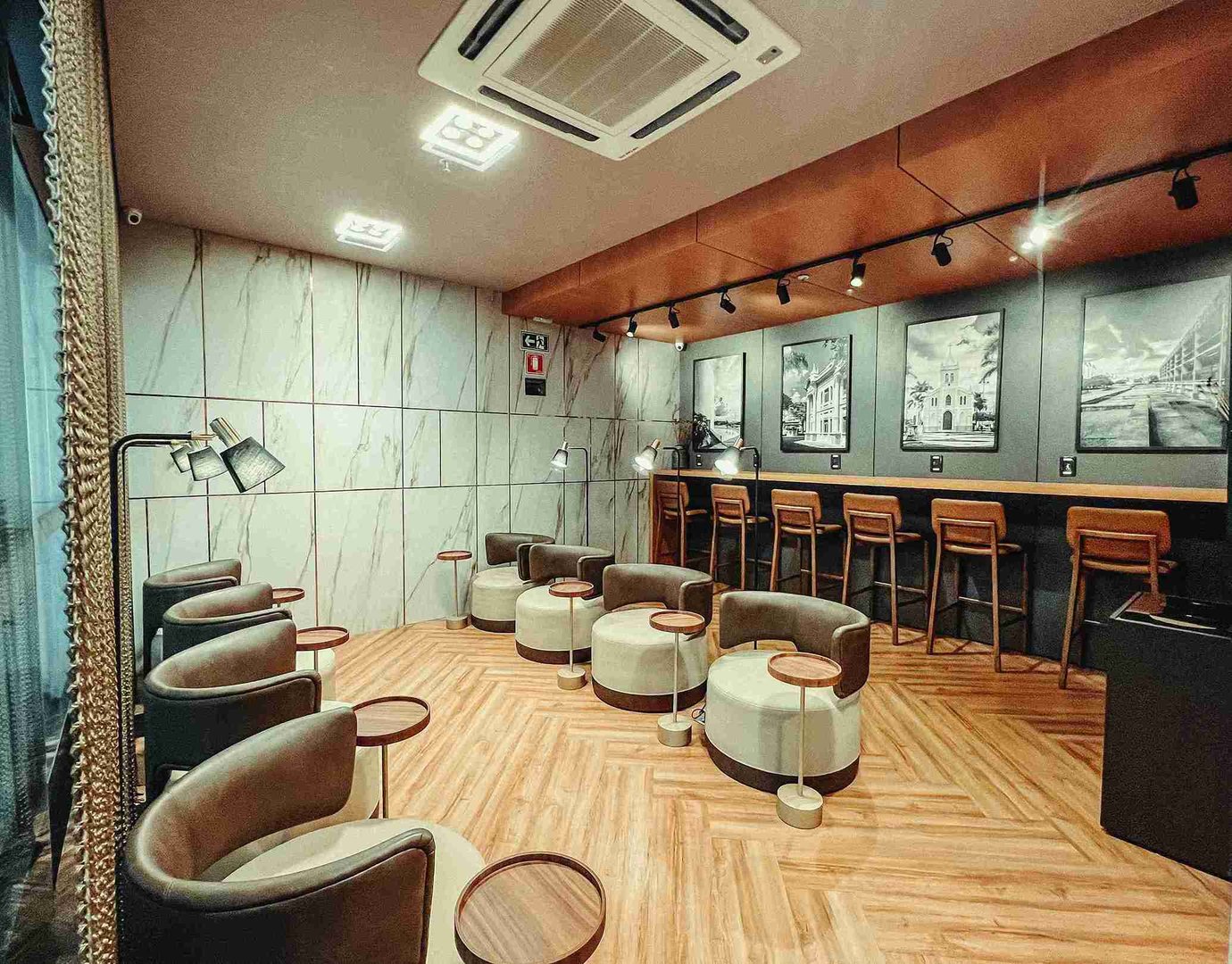 Quiet seating area in the W Lounge at Uberlândia Airport, featuring a row of rounded upholstered armchairs with small circular side tables on a wood-patterned floor, individual reading lamps, a marble-effect wall.