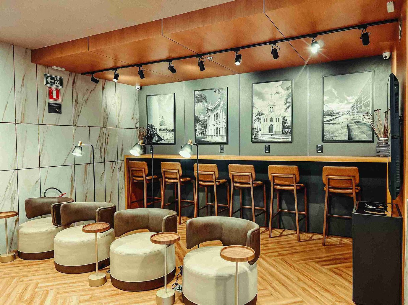 Bar-style seating area in the W Premium Lounge at Uberlândia Airport, with a long wooden counter lined with high stools, individual task lamps, framed black-and-white architectural photos on the wall, and a foreground of rounded lounge chairs with small circular side tables.
